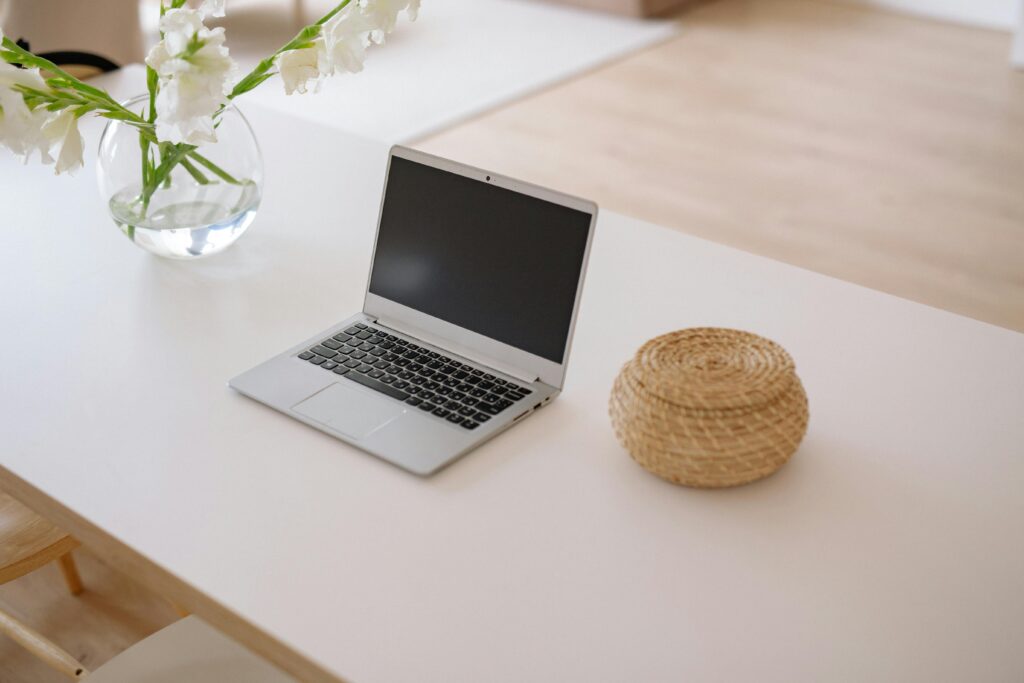 A clean and elegant workspace featuring a laptop, vase with white flowers, and a woven basket.